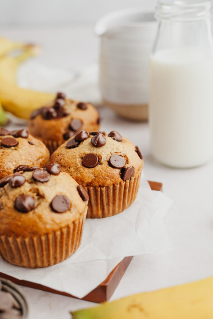 a side view photo of four banana muffins with chocolate chips in them on a cutting board lined with parchment paper next to a clear jug with milk in it