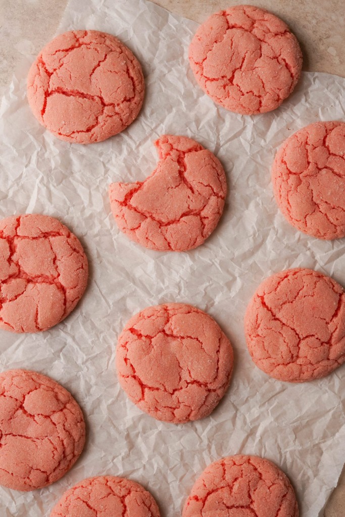 Above view of strawberry cookies with a crinkled top on a parchment line baking sheet. One of the strawberry cookies has a bite out of one of them.