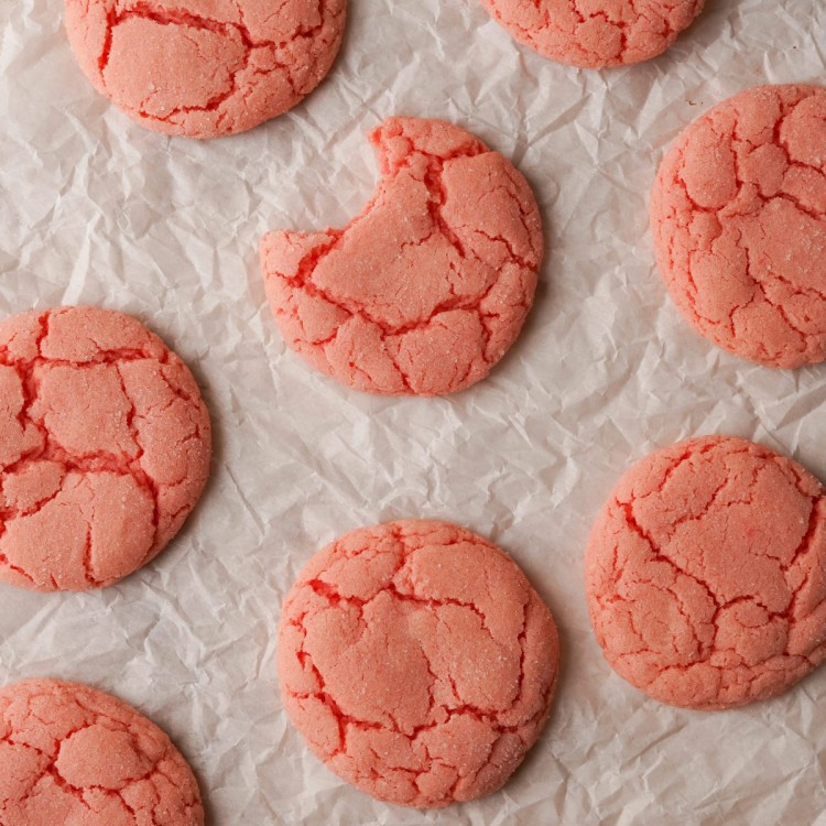 Above view of strawberry cookies with a crinkled top on a parchment line baking sheet. One of the strawberry cookies has a bite out of one of them.