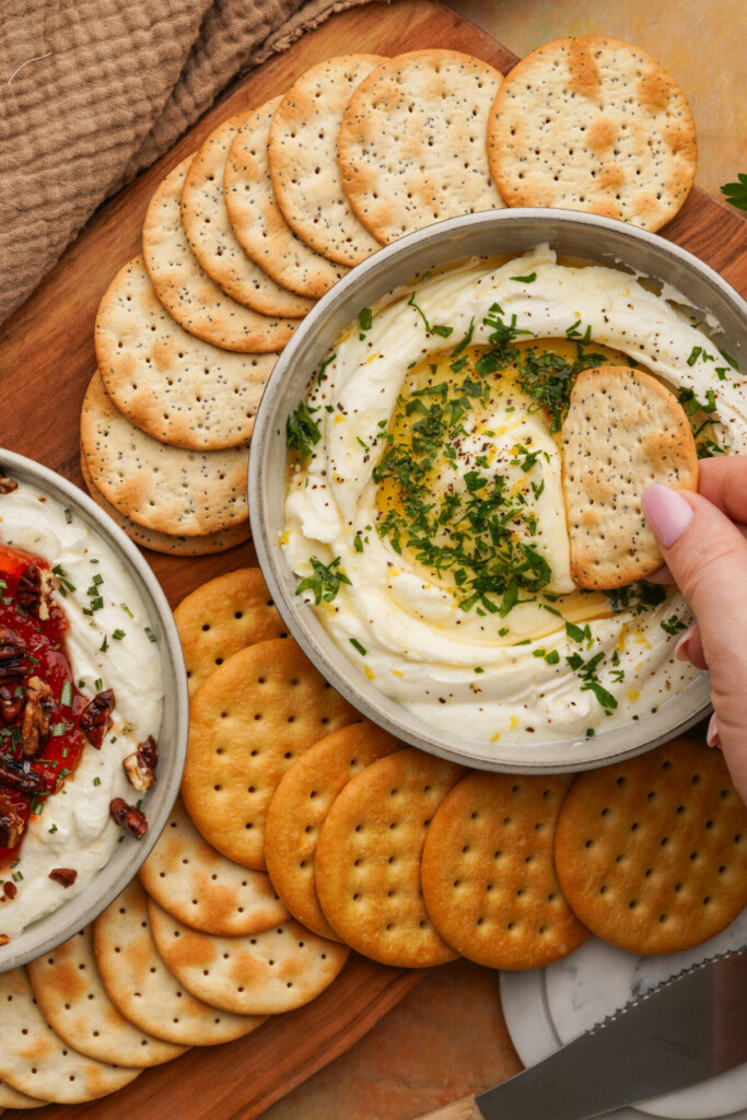 an above photo of goat cheese dip with crackers around it and a hand dipping a cracker in the dip