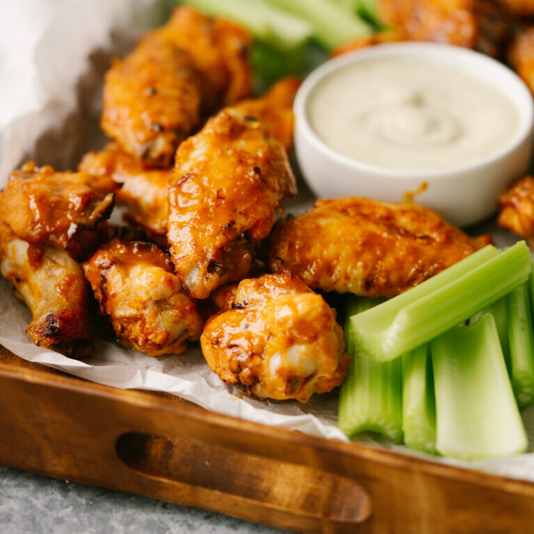 a photo of orange chicken wings on a wooden tray with a bowl of ranch and celery sticks