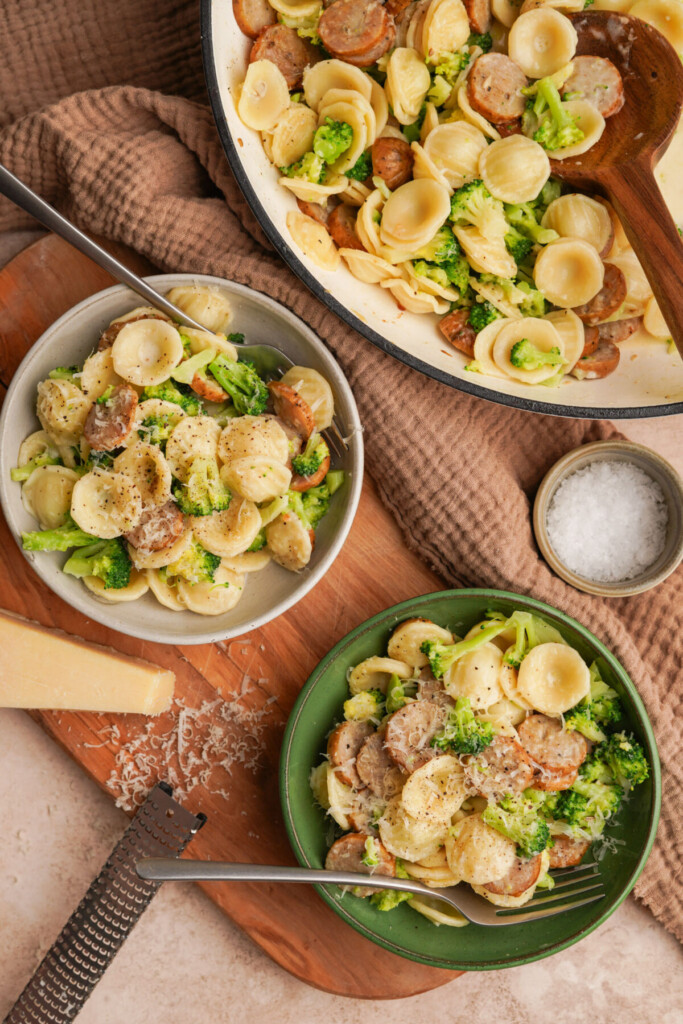 an above view photo of broccoli sausage pasta in a pot in the corner with two bowls of pasta and a block of parmesan cheese next to it