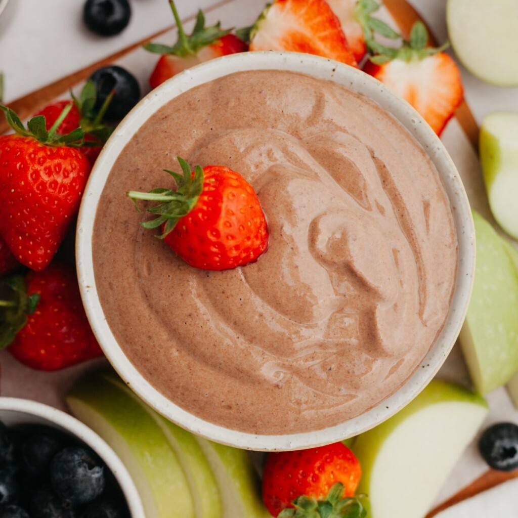 a photo of chocolate dip in a white bowl with a strawberry in it and fruit surrounding the bowl
