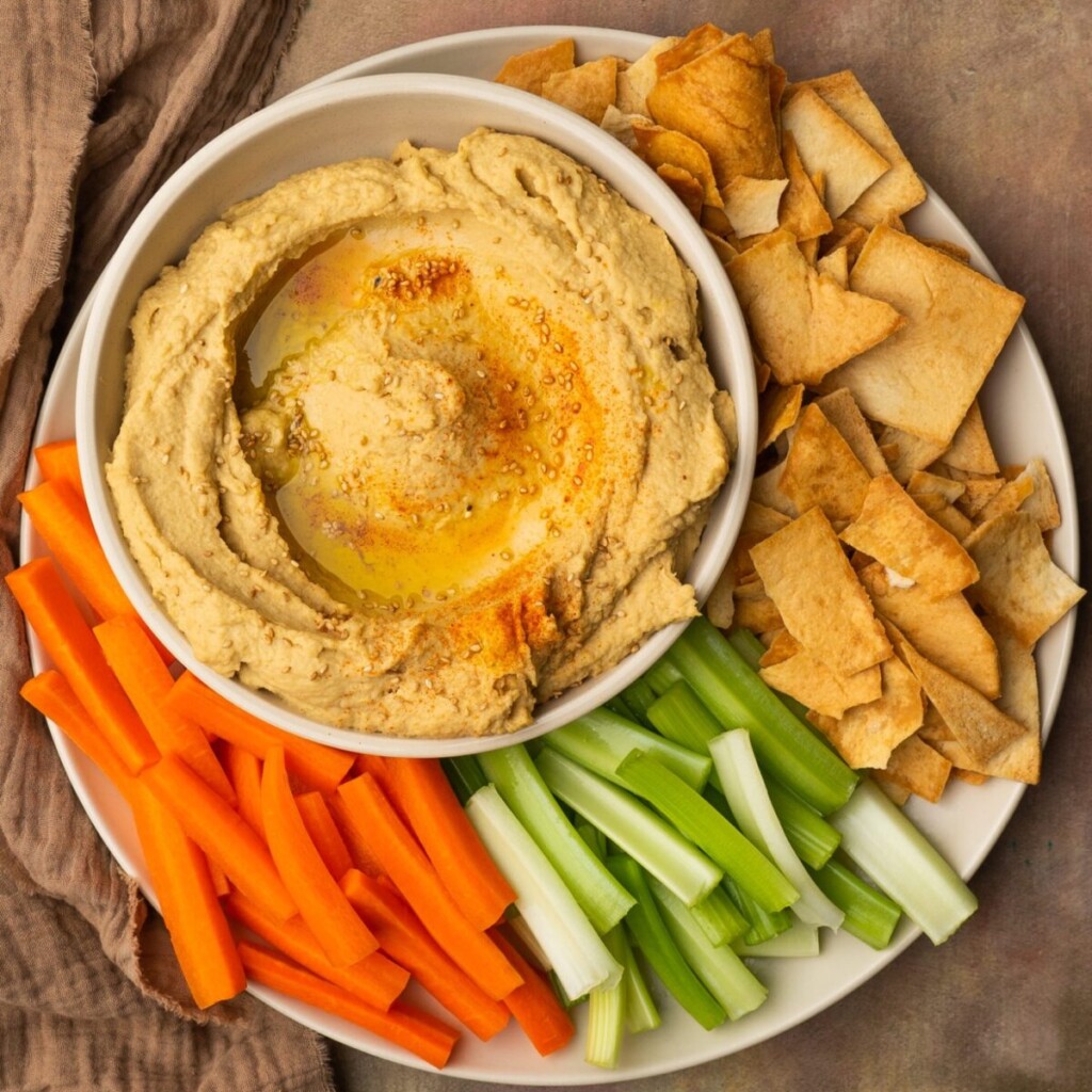 a photo of garlic hummus in a white bowl on a plate with carrots, celery, and pita chips