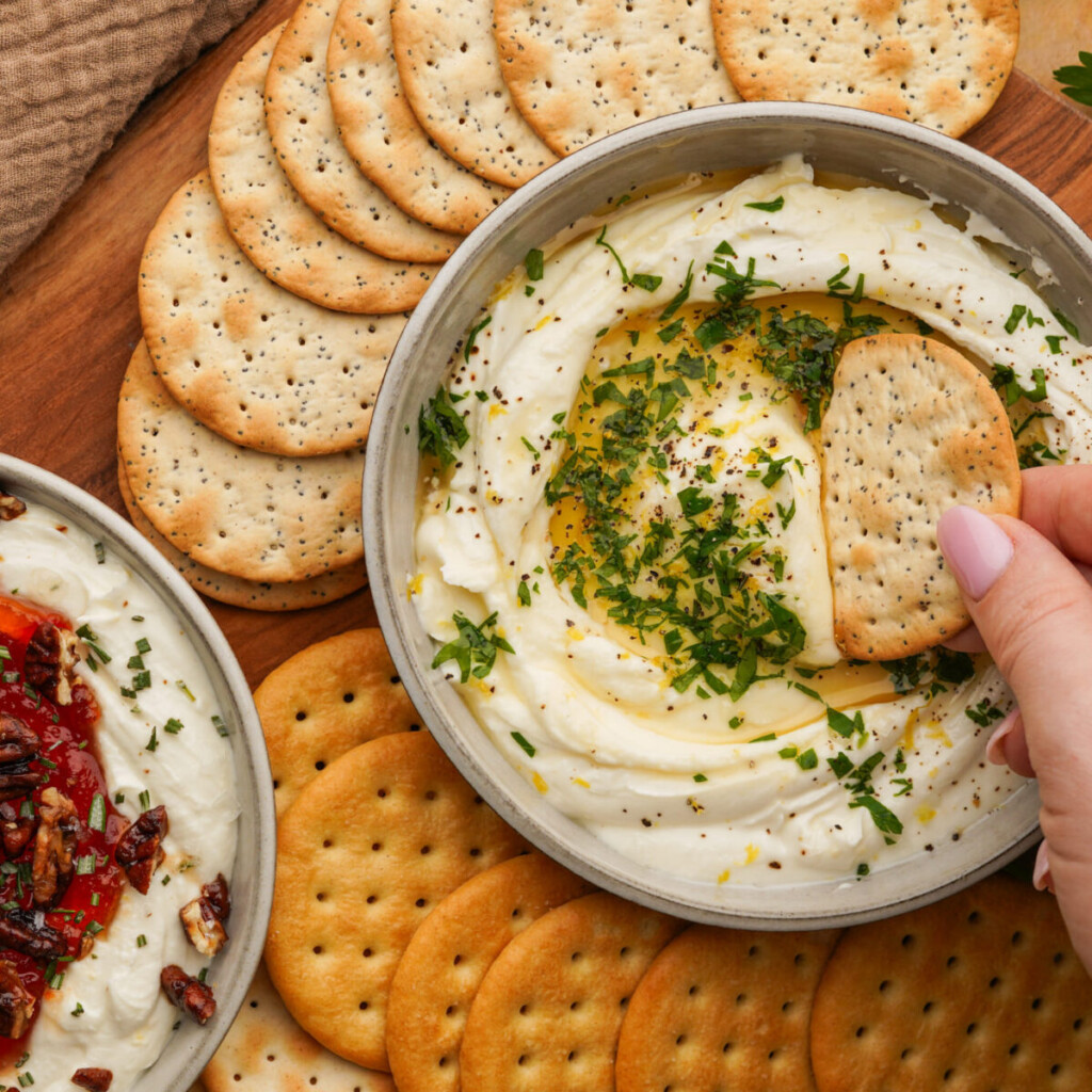 a photo of whipped goat cheese dip with a hand dipping a cracker in it and more crackers around it