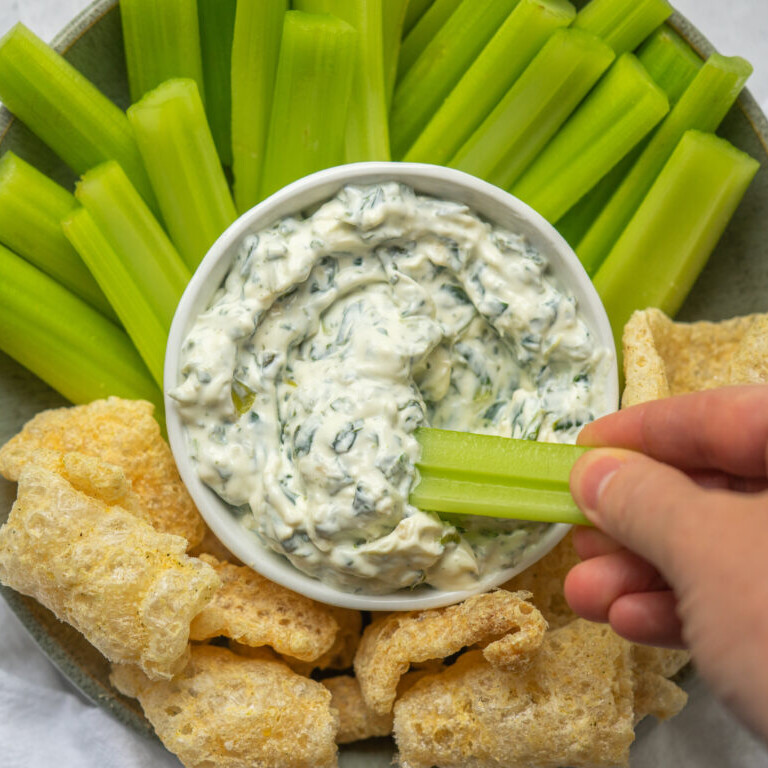 a photo of spinach dip with celery and chips around it and a hand dipping a celery stick into the dip
