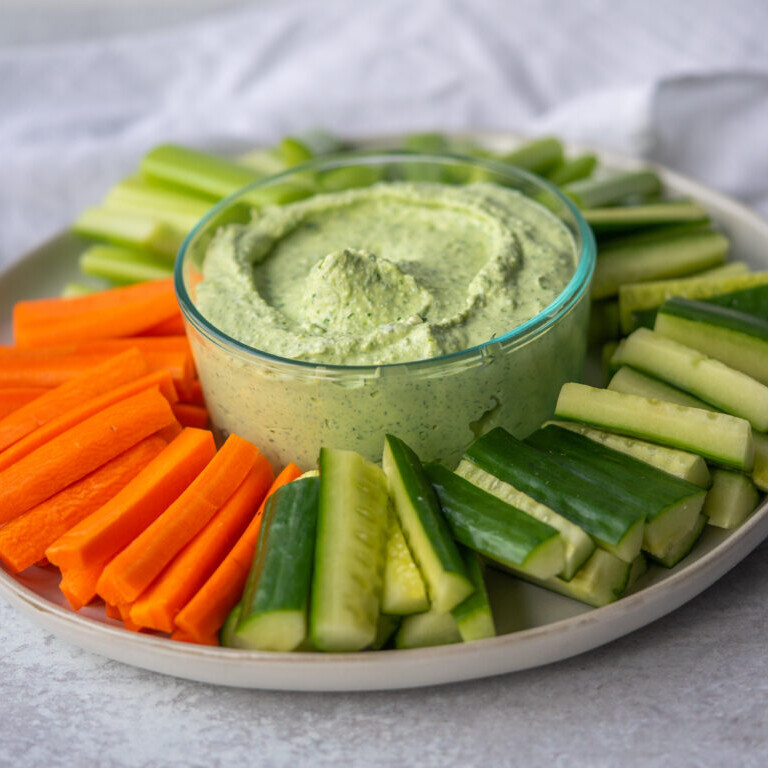 a photo of whipped feta dip in a clear bowl in the center of a white platter with carrot sticks, celery sticks, and cucumber sticks surrounding it