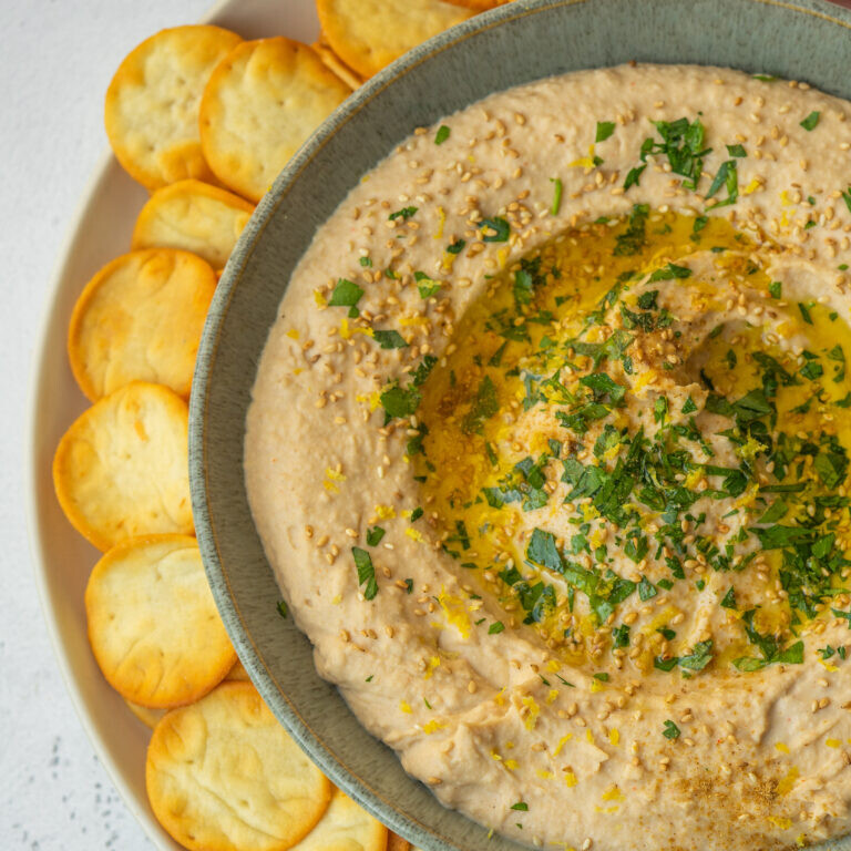 a photo of white bean hummus in a gray bowl with crackers around it