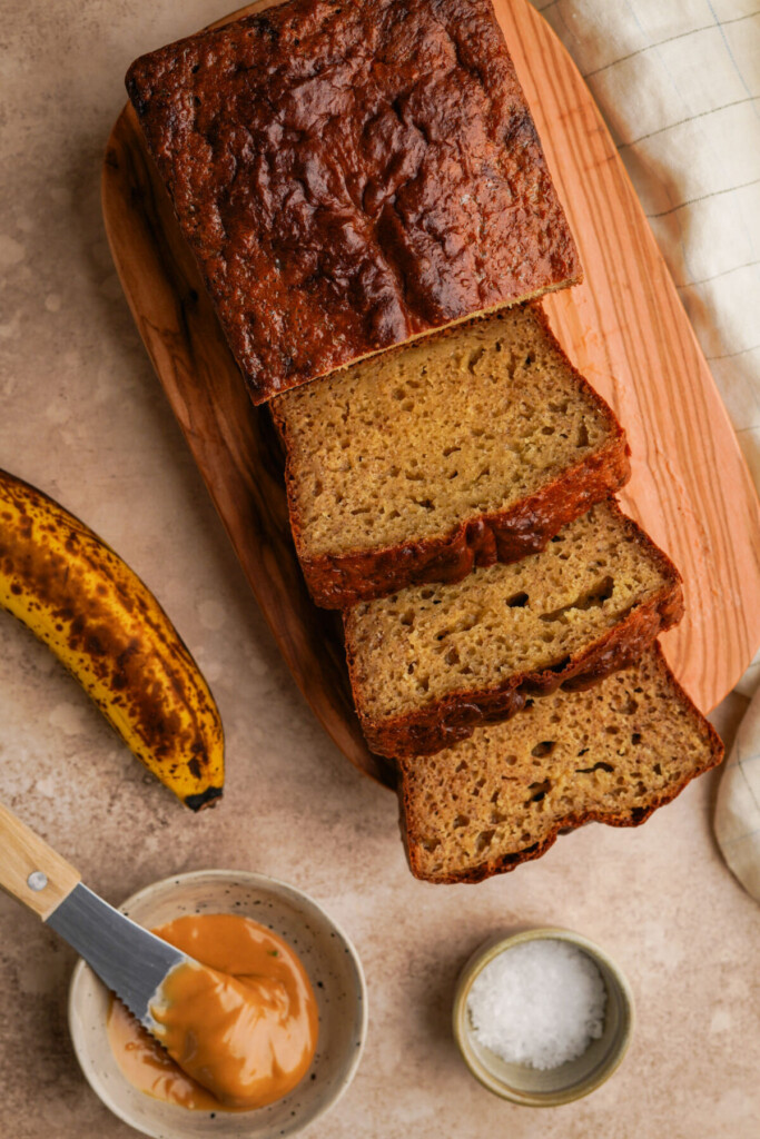 A loaf of protein banana bread cut into slices on a wooden cutting board next to a banana, peanut butter and salt