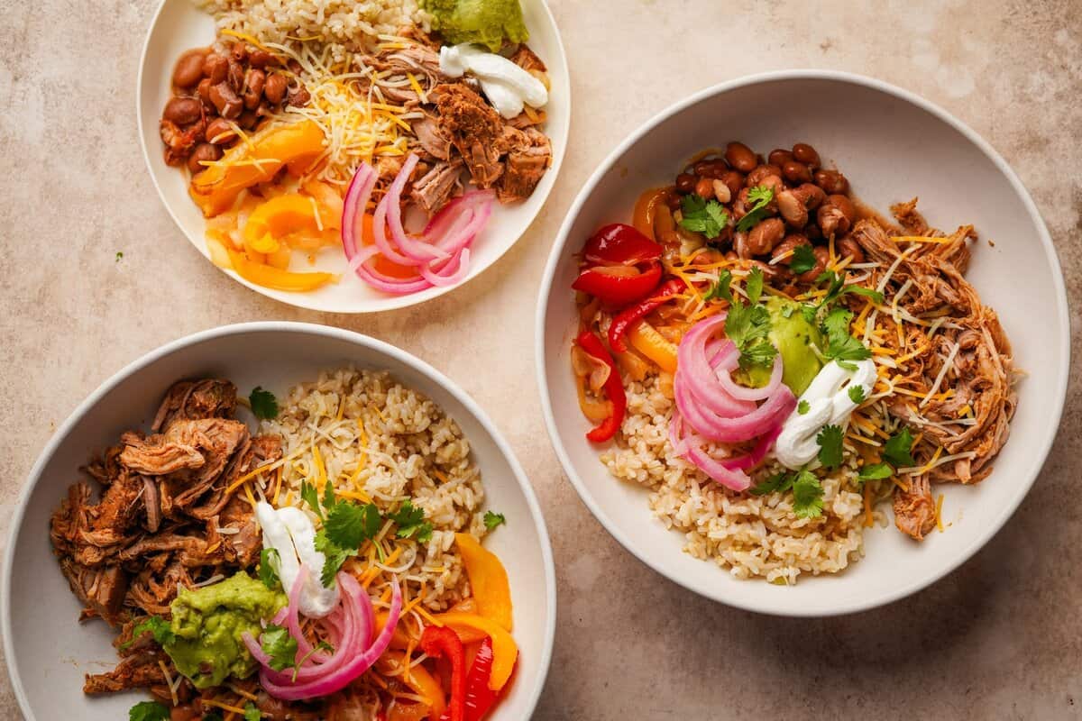 Serving bowls filled with rice, sauteed peppers and onions, shredded pork, beans and burrito bowl toppings