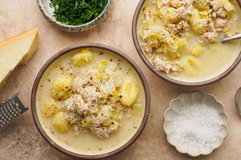 an above view image of two bowls of chicken leek soup with salt and parsley in small bowls around it
