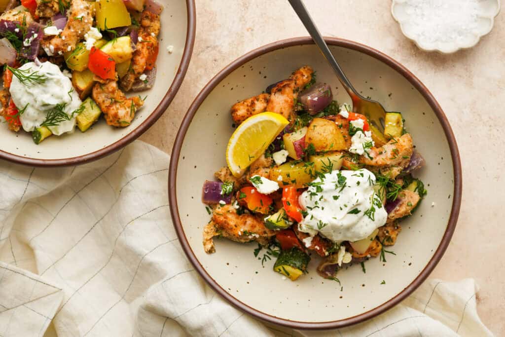 an above view photo of greek sheet pan chicken and veggies in a bowl with lemon and tzatziki on top