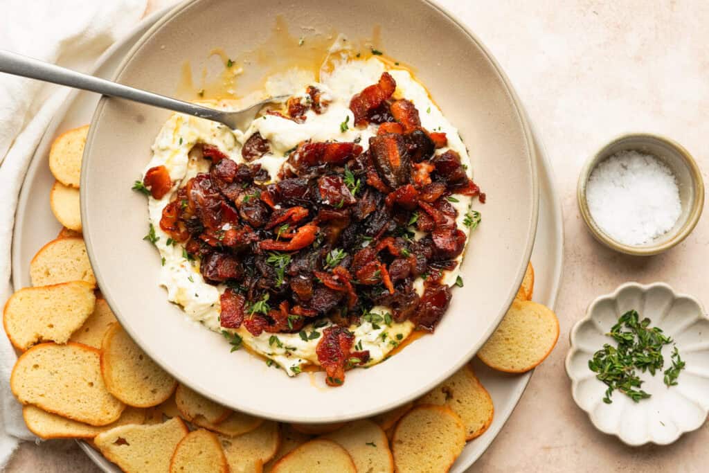Above view of sauteed bacon and dates on cheese in a bowl with some bagel chips around it and fresh herbs on top
