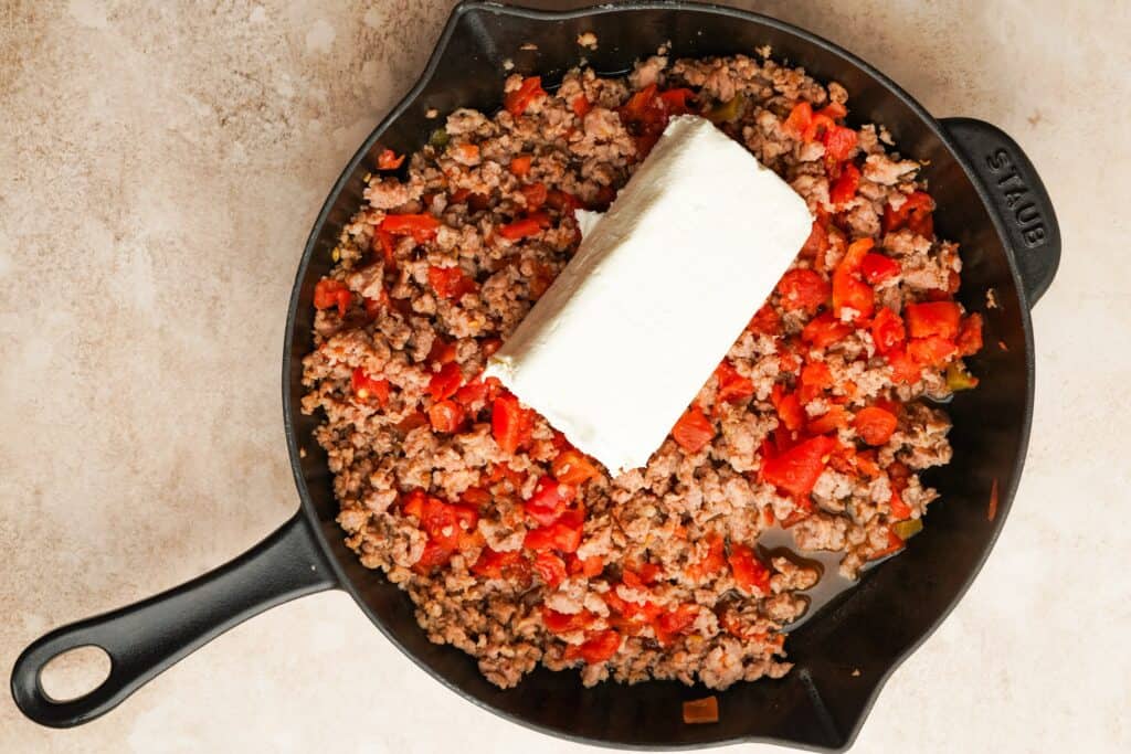 Cooked ground sausage, and a can of rotel in a cast iron skillet with a brick of cream cheese in the middle.
