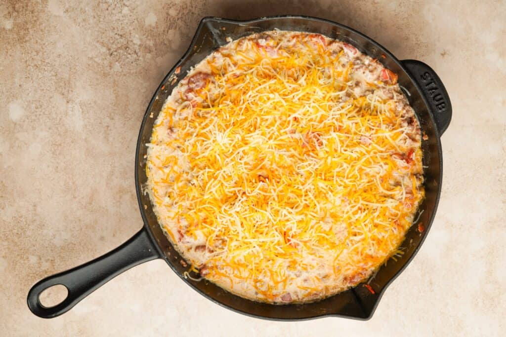 Ground sausage, rotel and cream cheese mixed together in a cast iron skillet with shredded cheddar cheese on top.