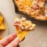 A hand holding a tortilla chip that has cheesy sausage rotel dip on it