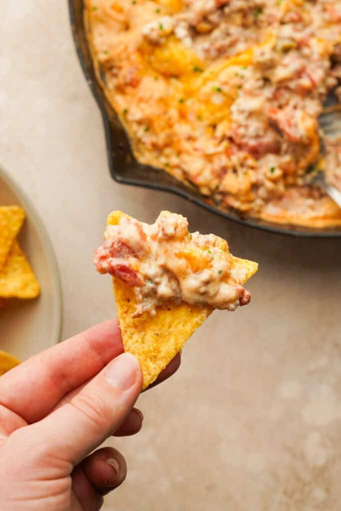 A hand holding a tortilla chip that has cheesy sausage rotel dip on it