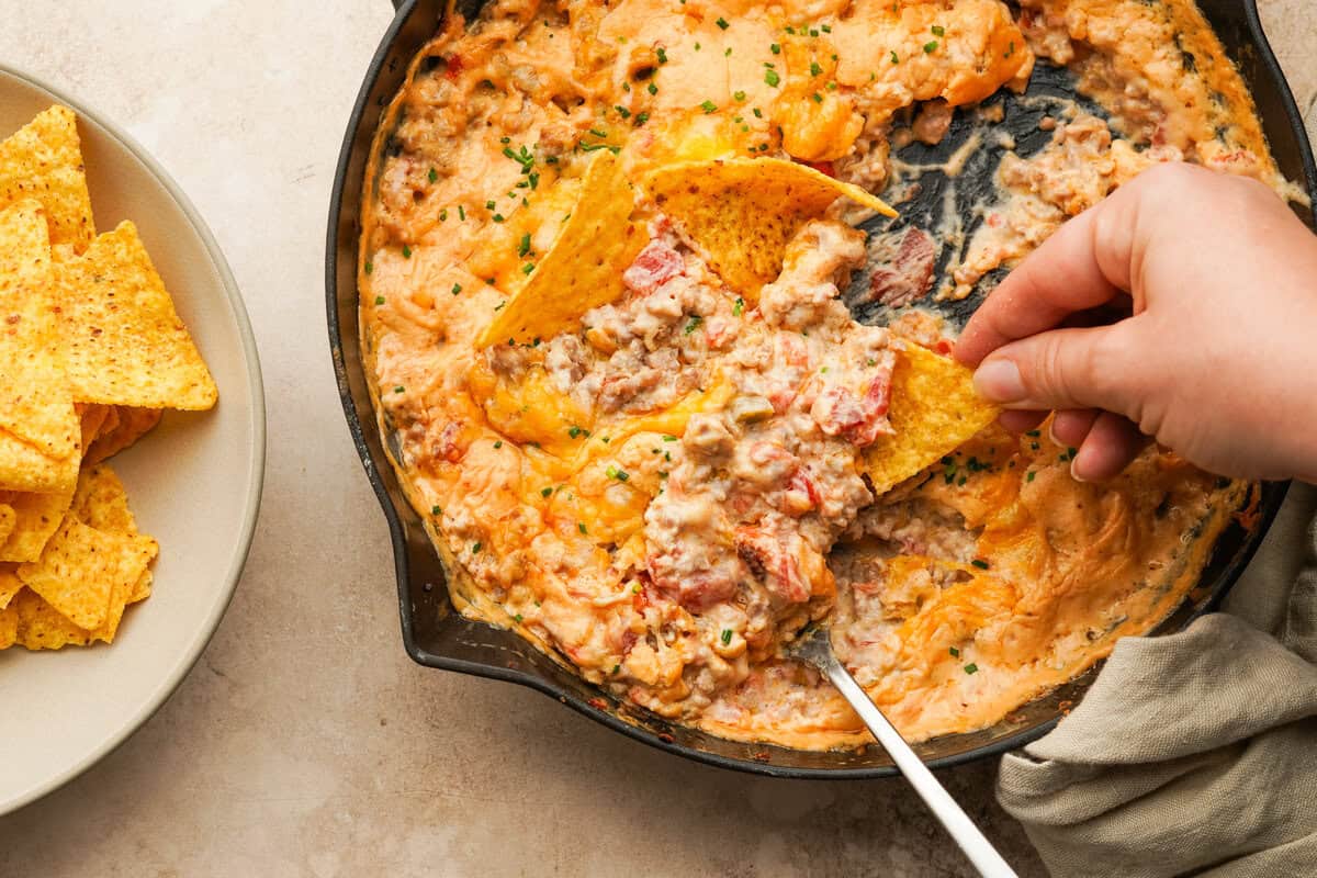 A hand holding a chip dipping into a cheesy rotel sausage dip in a cast iron skillet