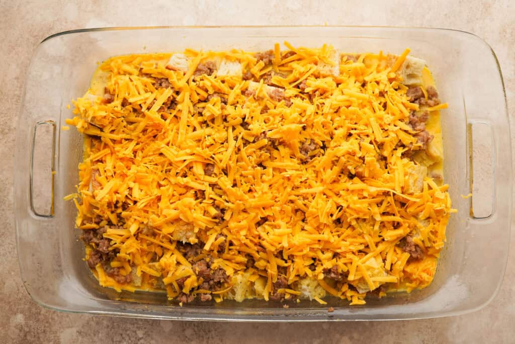 A casserole dish filled with cubed bread, breakfast sausage, egg and milk mixture and shredded cheddar cheese