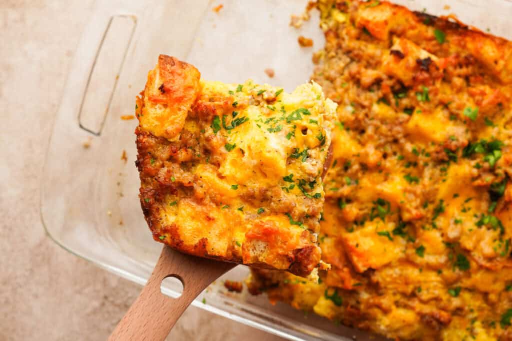 A spatula holding a cheesy slice of sausage strata above a casserole dish