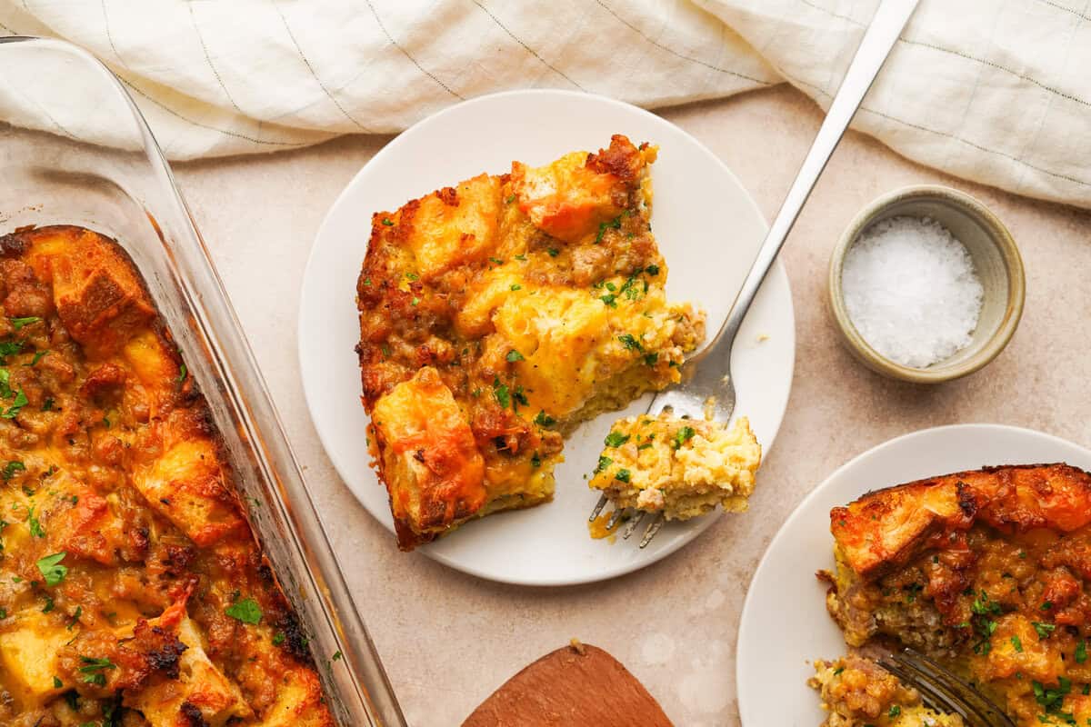 A serving plate with a slice of sausage strata breakfast casserole on it and a fork cutting into the casserole