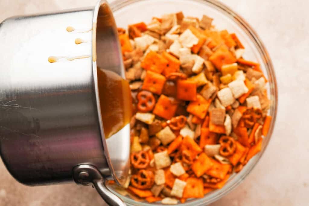 Sugar sauce pouring out of a pot into the bowl with the chex, pretzels, peanuts and cheez its
