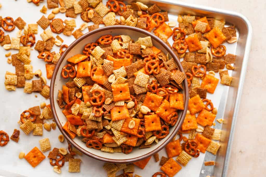 A serving bowl filled with sweet and salty chex mix sitting on a baking sheet with some of the chex mix on it