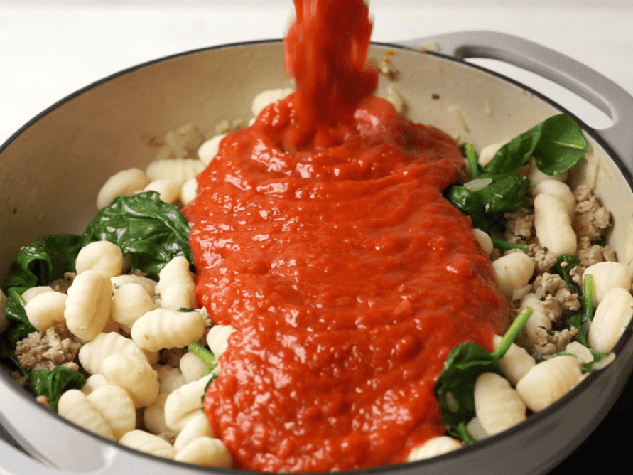 Marinara sauce being poured into a large skillet with gnocchi, ground turkey spinach and yellow onions.