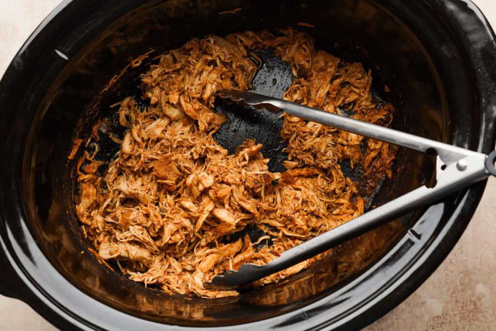 overhead shot of a crockpot with shredded chicken and tongs in it