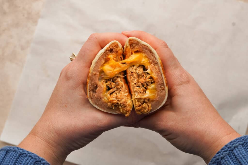 two hands holding a cut open chicken burrito with cheese oozing out
