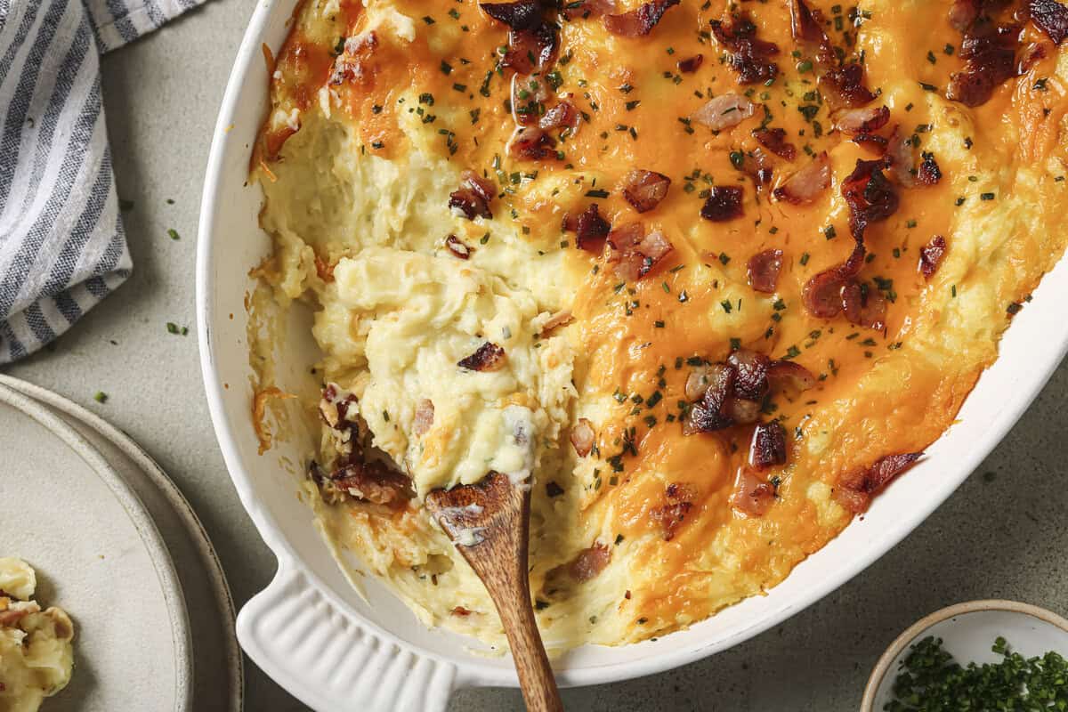 overhead shot of a baked potato casserole with a piece missing, serving plates and a side of herbs are shown off to the side 