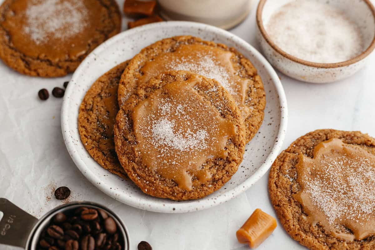 caramel latte cookies on a serving plate, with sugar, espresso beans, and a caramel candy in background