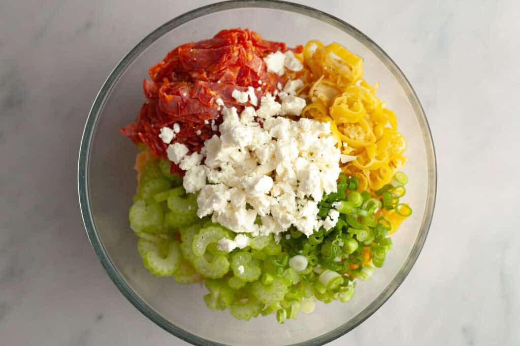 sliced pepperoni, pepperoncini, celery, crumbled feta and green onion in a mixing bowl on top of cooked pasta