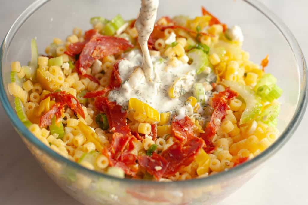Pepperoncini sauce being poured into a mixing bowl with pasta, pepperoni, sliced pepperoncini, celery, and green onion