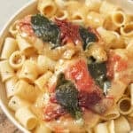 chicken saltimbocca on top of rigatoni pasta in a white bowl on a wooden cutting board