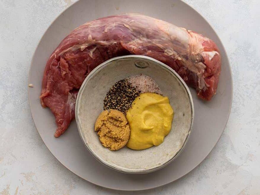pork tenderloin on a plate next to a small bowl with brown sugar, dijon mustard, salt and pepper