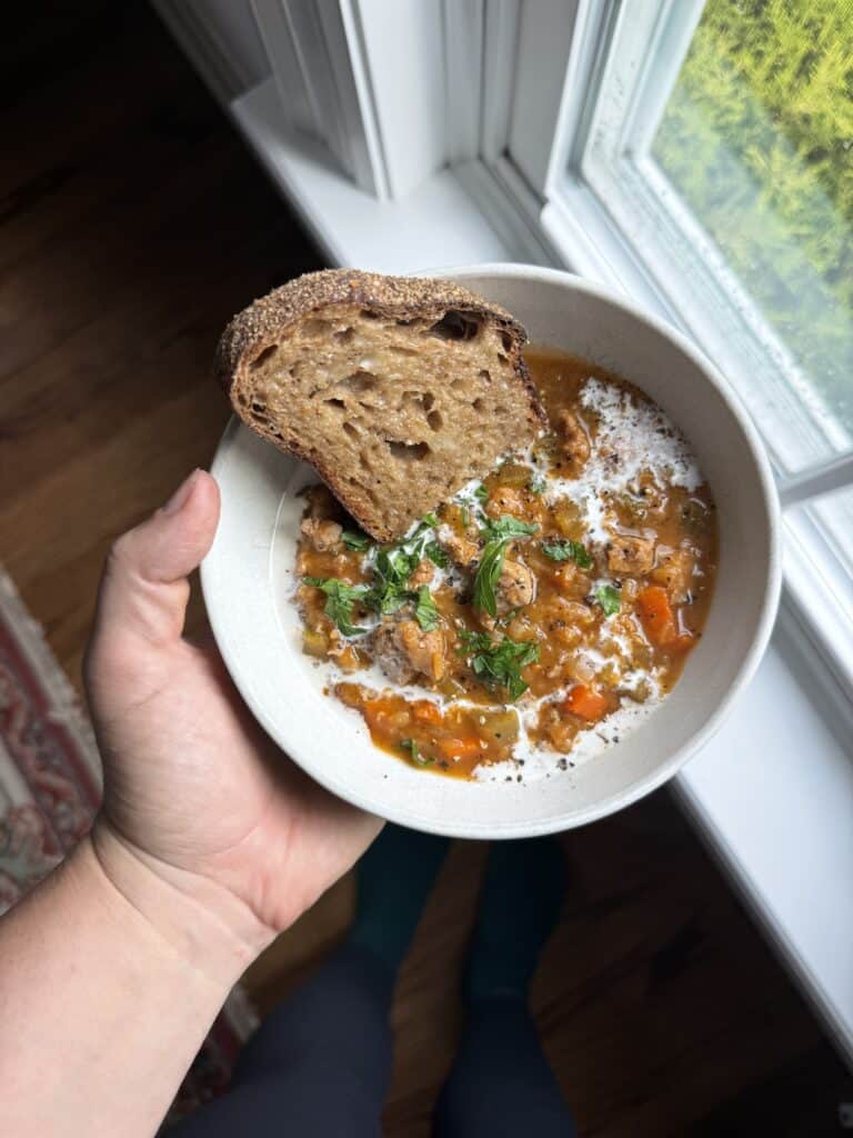 a hand holding a bowl of lentil and sausage soup with a drizzle of heavy cream and fresh herbs on the soup and slice of brown sourdough bread in the bowl