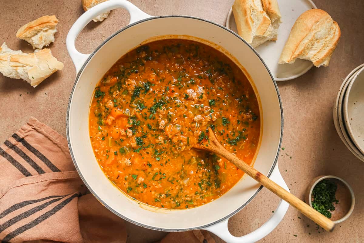 lentil and sausage soup made with creamy red lentils in a large pot with a wooden spoon in the pot. the pot is surrounded by a crusty baguette and some herbs